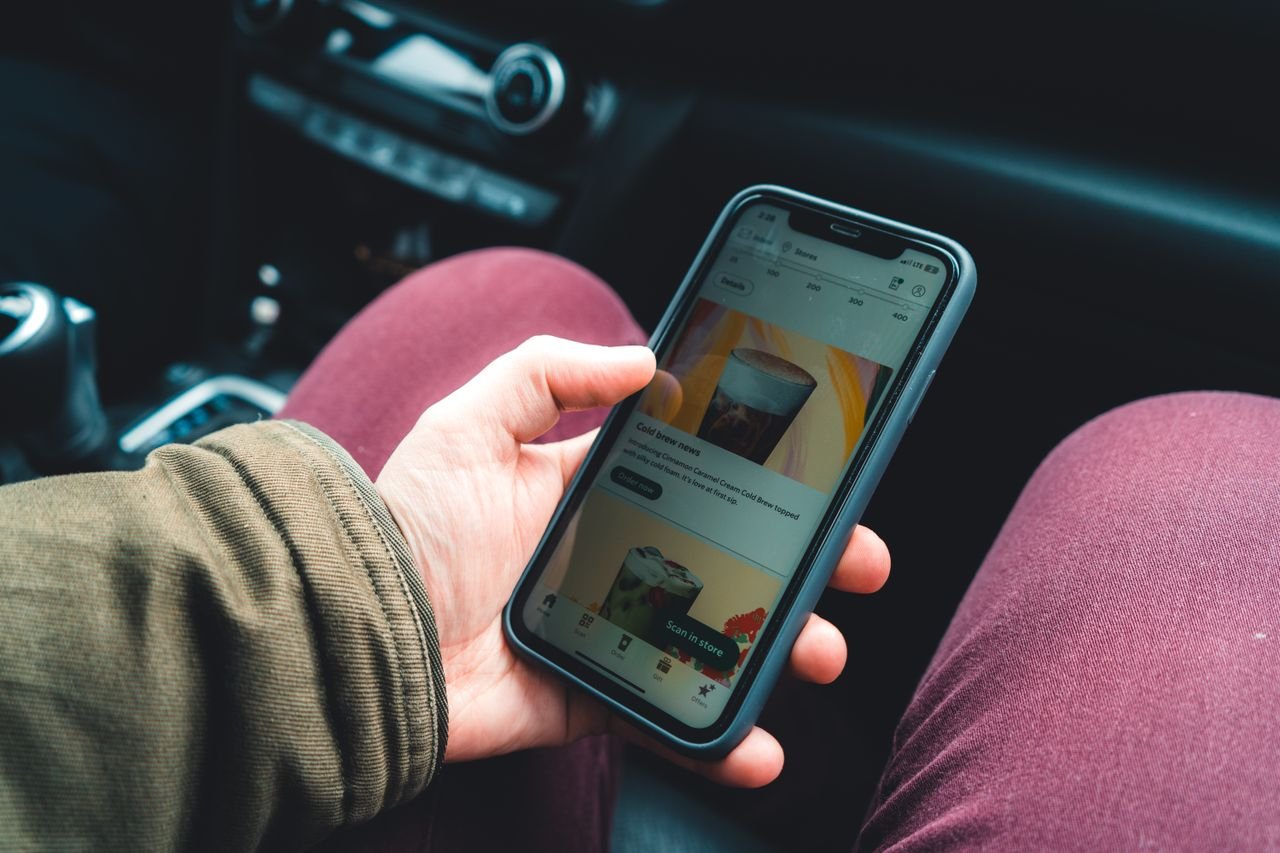 Hand holding smartphone showing a coffee app interface in a car interior setting.